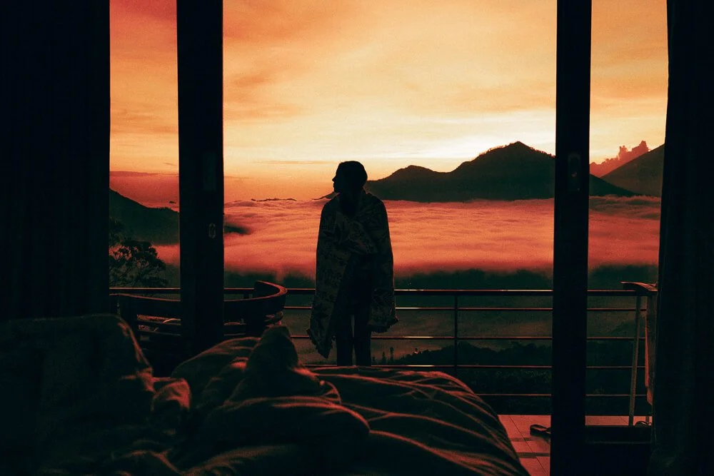 Person standing on a balcony, overlooking a valley with clouds and mountains during sunset, seen from inside a room.
