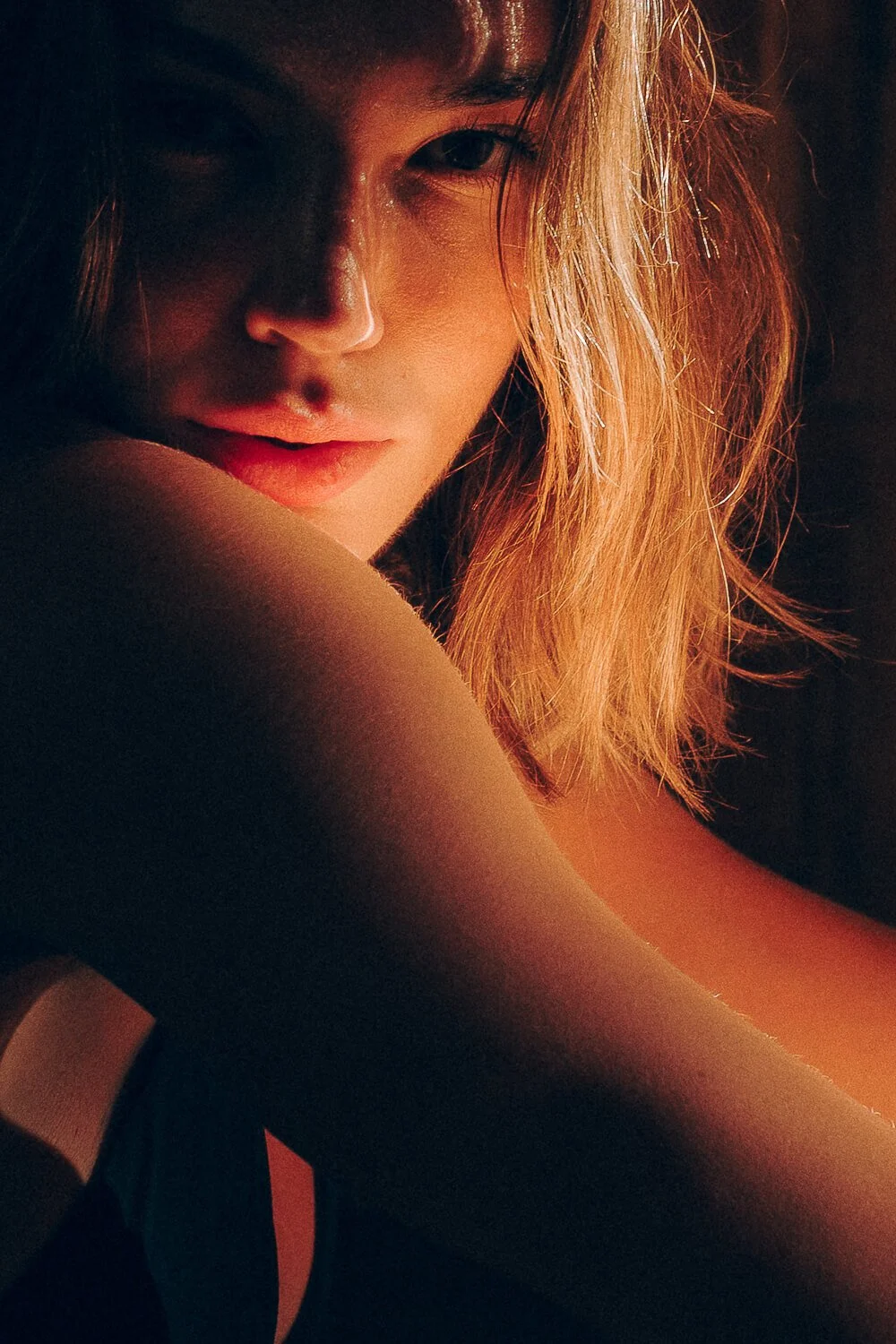 Close-up of a woman with long, wavy blonde hair, partially obscured face, soft lighting highlighting her features, against a dark background.