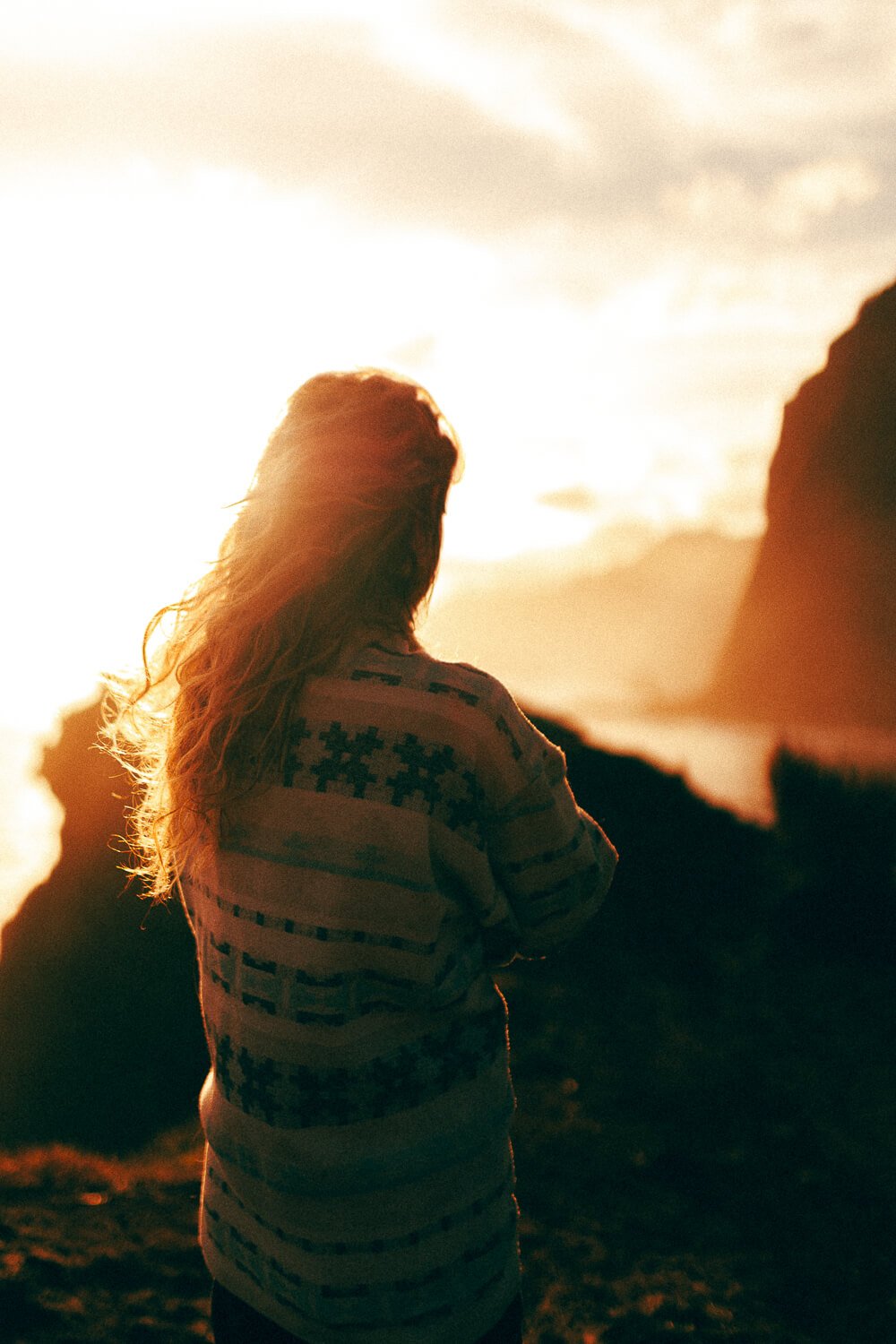 A woman with long hair standing outdoors at sunset, facing away from the camera, wearing a patterned sweater.