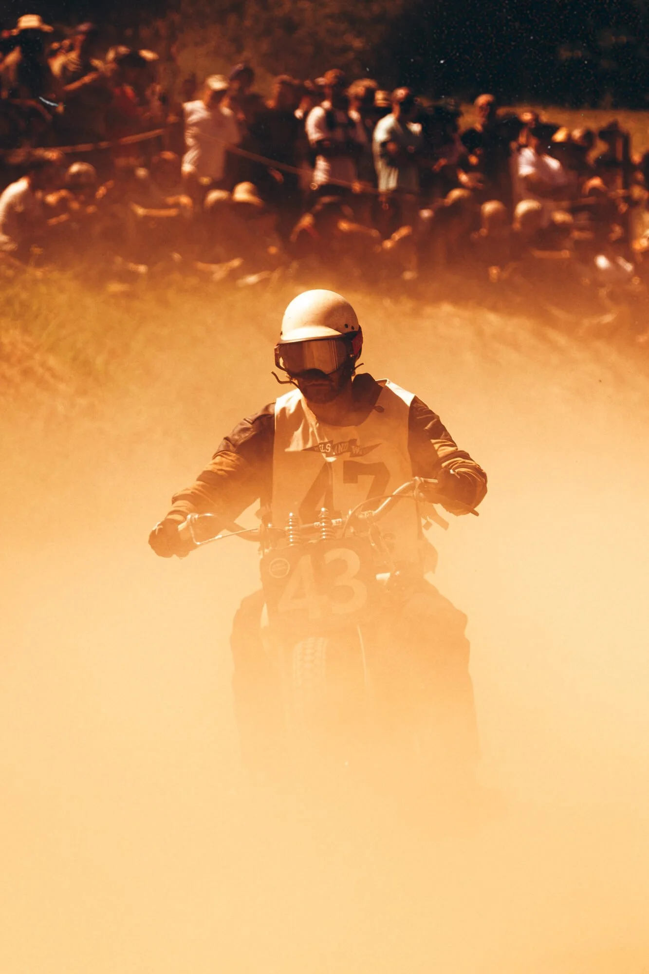 A person wearing a helmet and goggles riding a motorcycle through dust, with a crowd of spectators in the background.