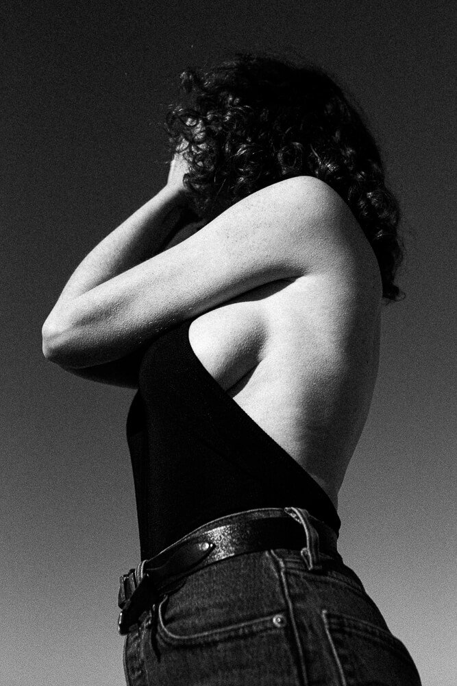 A woman with curly hair seen from the back, wearing a sleeveless top and jeans, with her arm across her face, against a clear sky.