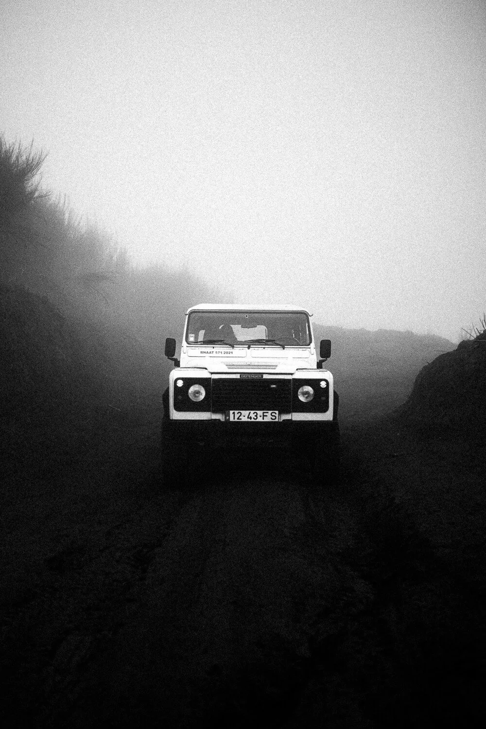 A black and white photo of a Land Rover seen from the front, driving on a misty, rough dirt road surrounded by hills.