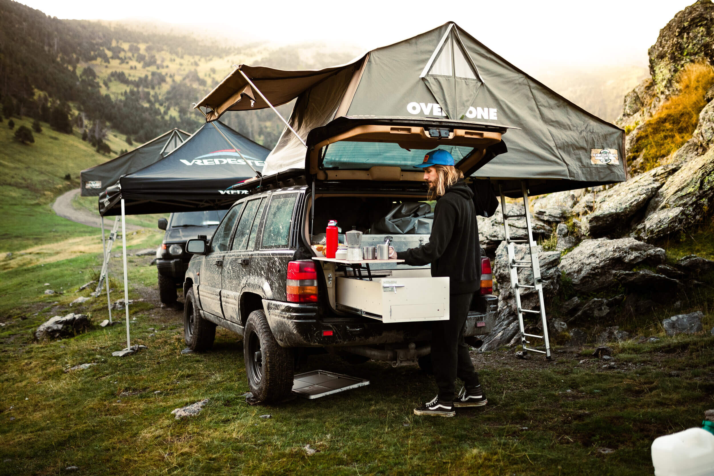 Un véhicule tout-terrain avec une tente de toit déployée. Une personne prépare du café à l'arrière du véhicule dans un cadre de camping en montagne.