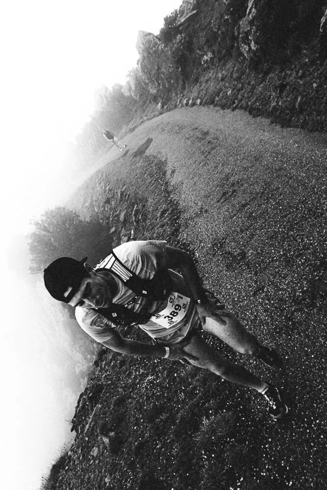 A person running on a rugged outdoor trail surrounded by trees in a mountainous area, wearing a race bib and a cap, captured in black and white.