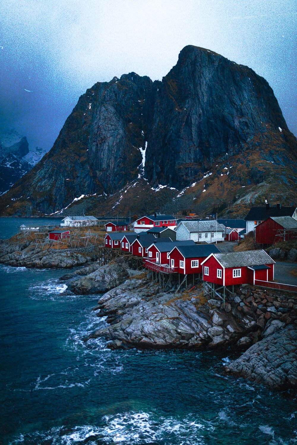 Un petit village de maisons rouges en bois sur la côte rocheuse, avec une grande montagne en arrière-plan et une mer agitée, dans un paysage scandinave.