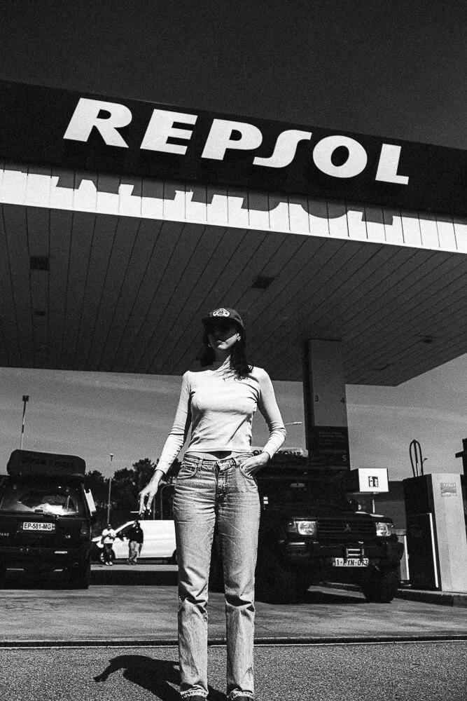 A woman standing at a gas station under a large Repsol sign, with cars and people in the background, in black and white.