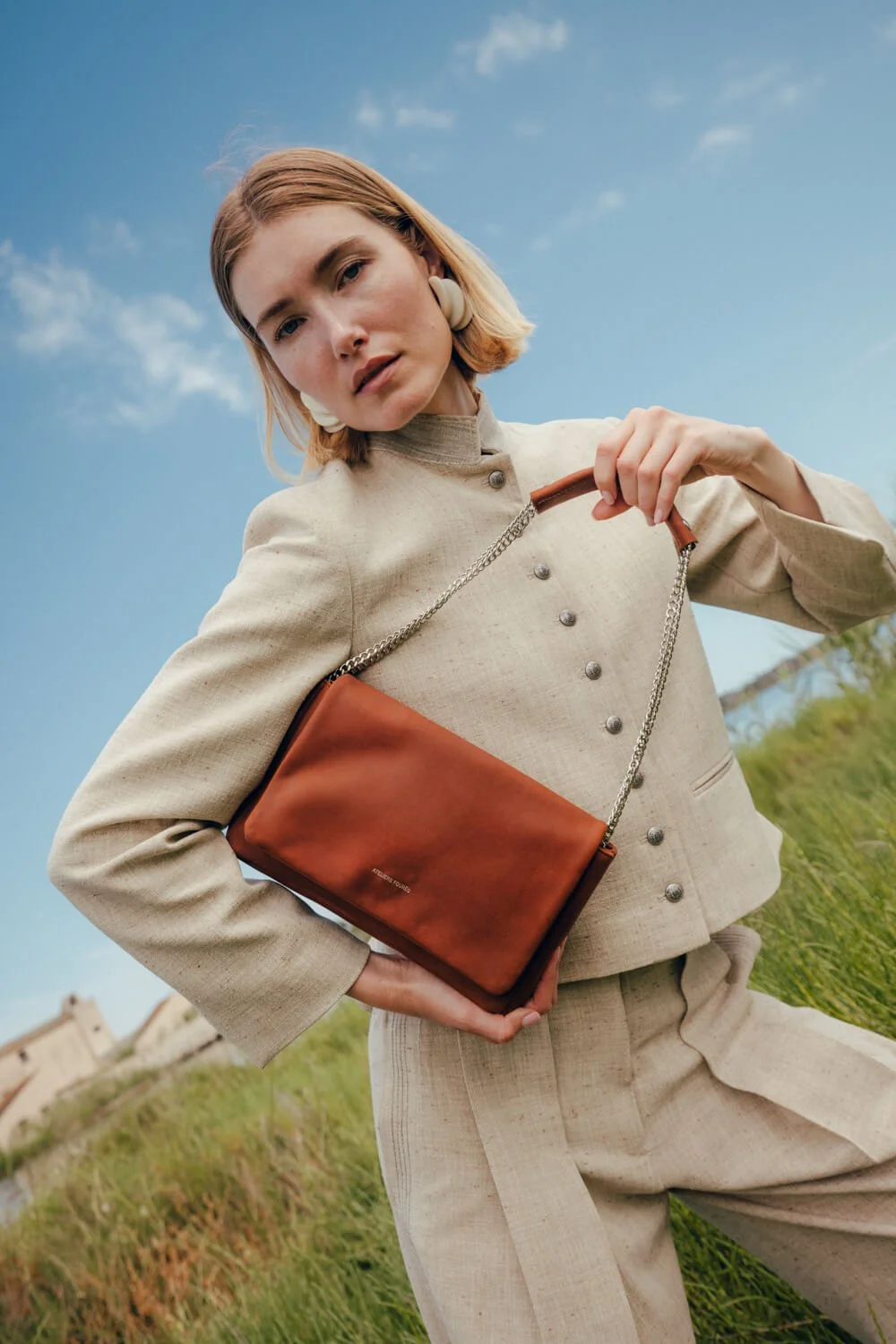 A woman with blonde hair wearing a beige two-piece suit with metallic buttons, holding a brown handbag with a chain strap outdoors against a blue sky with some clouds.