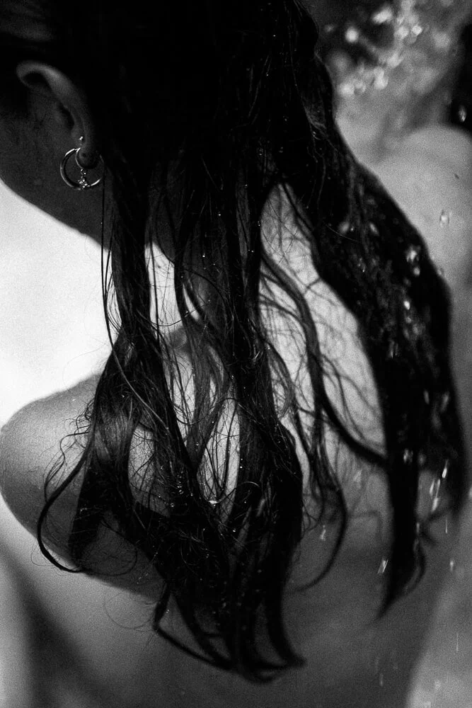 A person with wet, dark hair and hoop earrings, partly visible, sitting or leaning in a setting with water droplets, captured in black and white.
