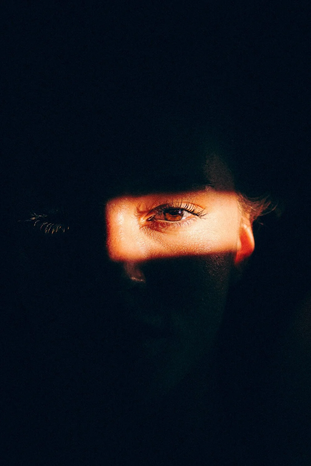 Partial view of a person's face with one eye visible through a horizontal cutout, surrounded by darkness.