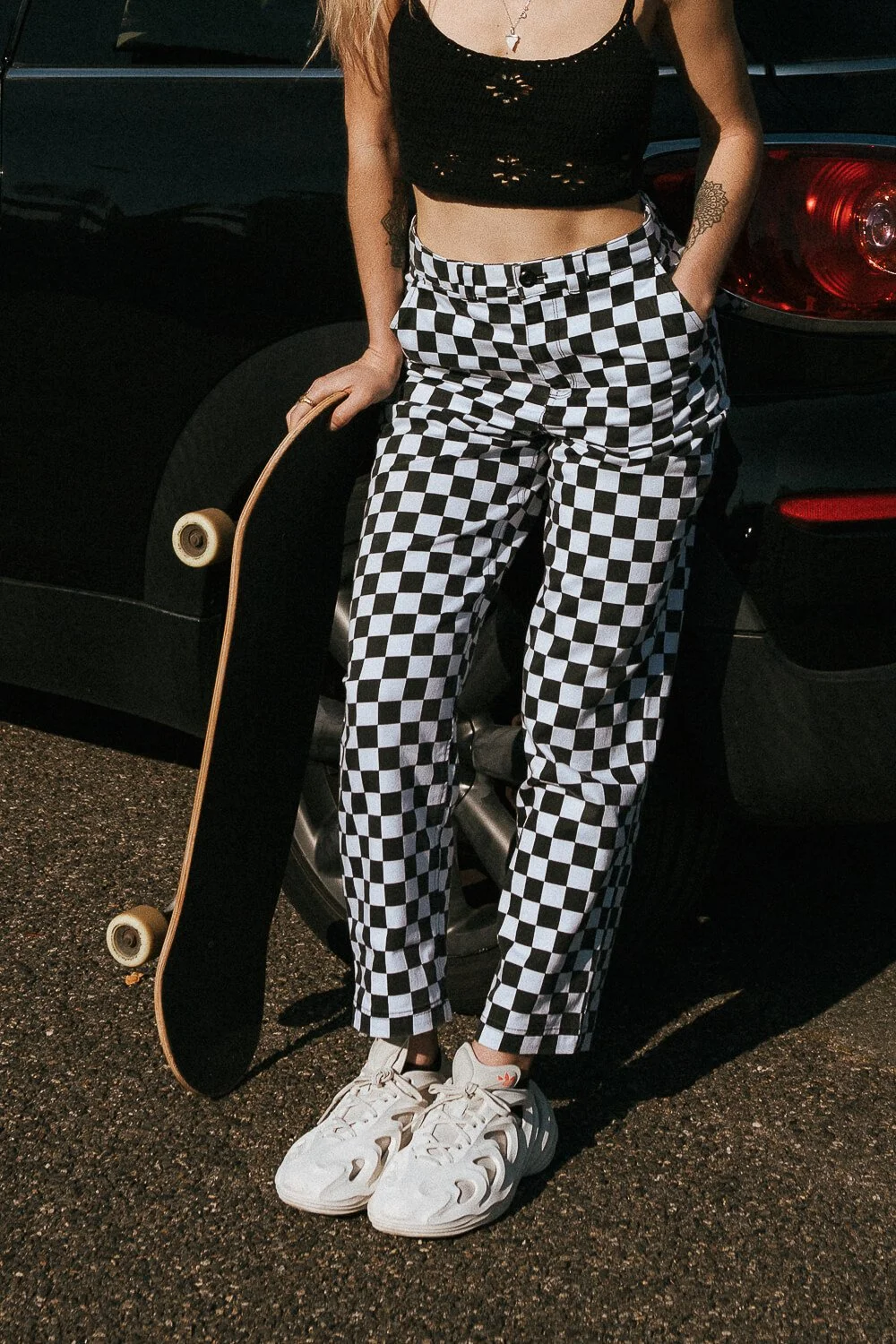 Person wearing a black crop top with holes, checkered black and white pants, and white sneakers, holding a skateboard, standing next to a black car.