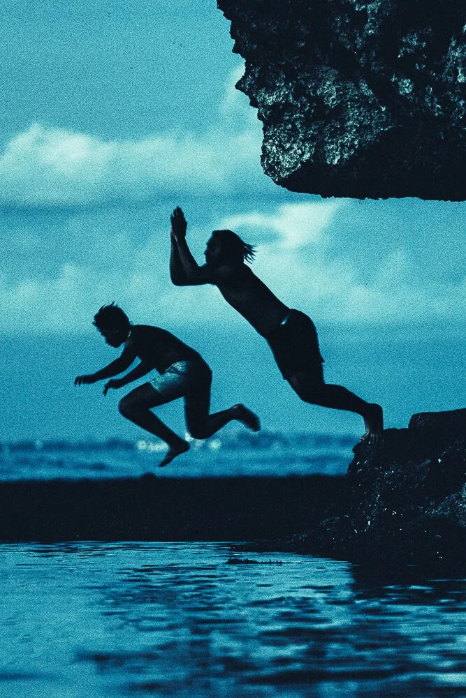 Two people jumping off rocks into water at the beach during sunset or dusk, silhouetted against the sky.