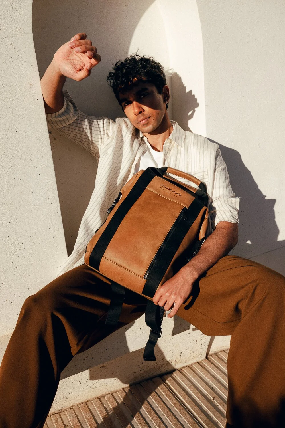 A young man with curly hair sitting against a white wall, holding a tan and black bag, wearing a cream-striped shirt and brown pants, in warm sunlight.