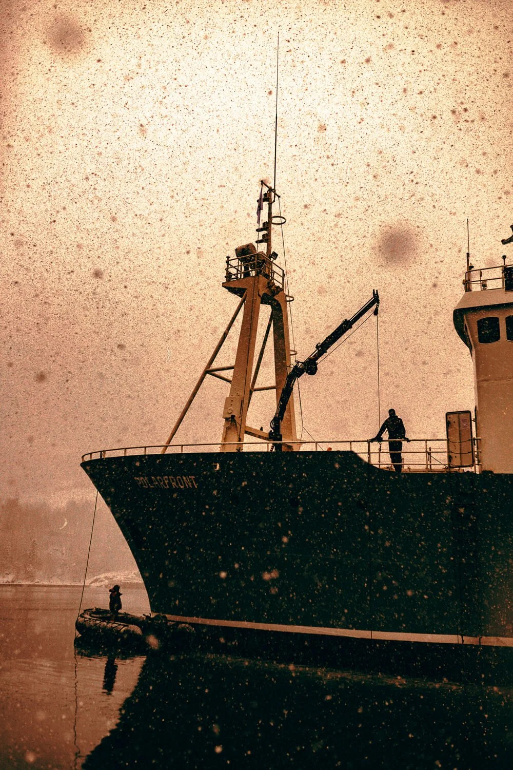 Un navire de pêche nommé 'POLARFRONT' en mer, avec deux personnes visibles sur le pont, un sur le pont principal et une autre sur une petite embarcation, sous un ciel brumeux et poussiéreux.