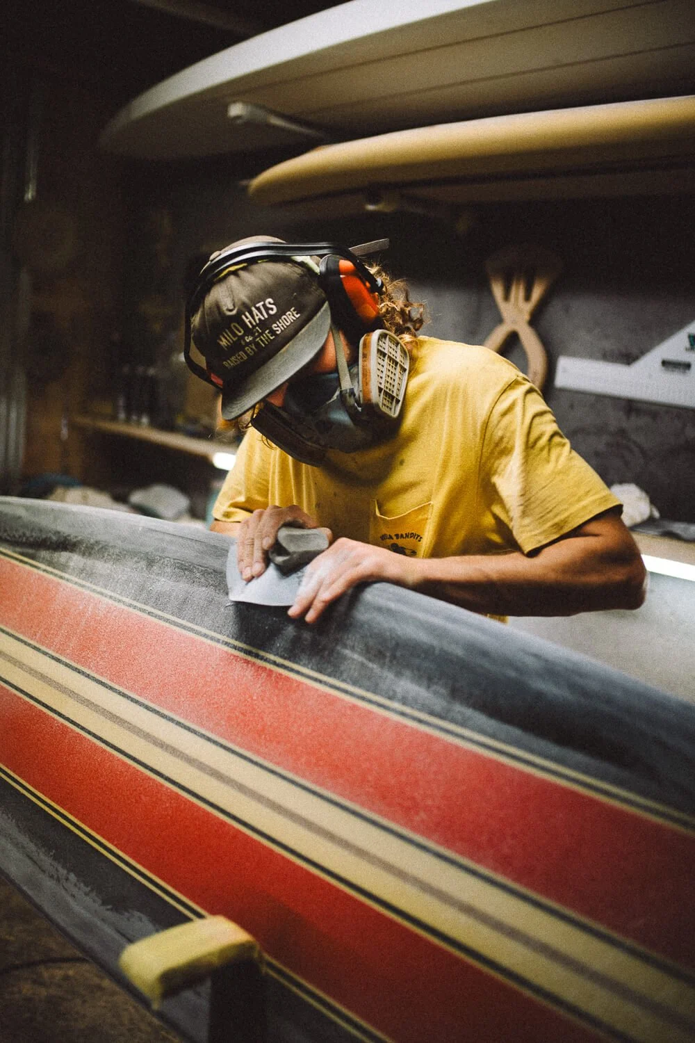 A person wearing a yellow shirt, riding a black and red surfboard, with a protective mask and noise-canceling headphones, sanding or polishing the surfboard inside a workshop.