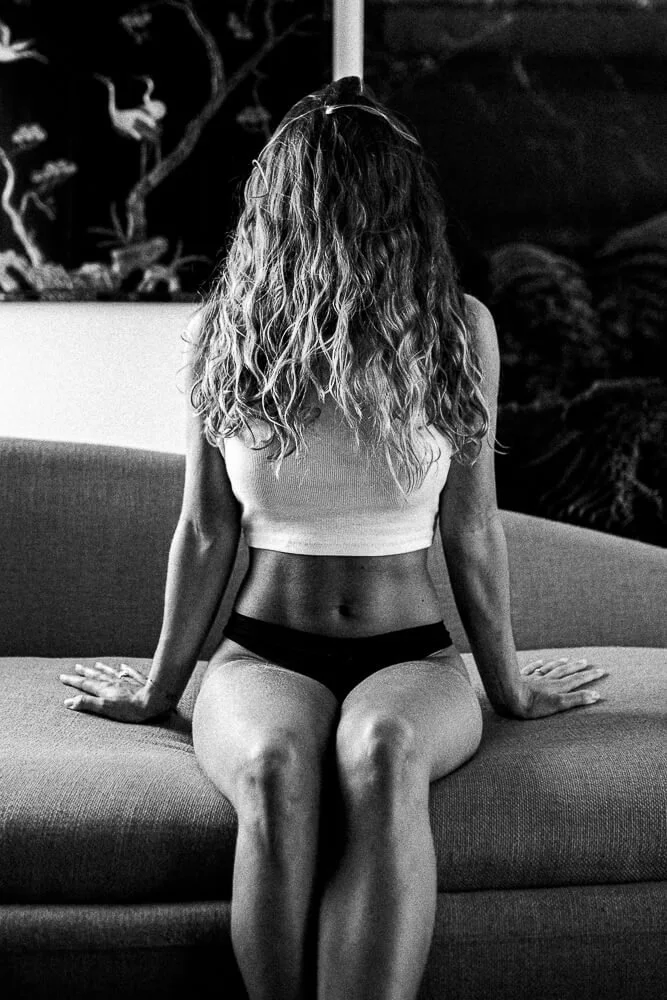 A woman with long curly hair sitting on a couch, facing away, wearing a sleeveless top and black underwear, with her hands resting on the couch beside her.