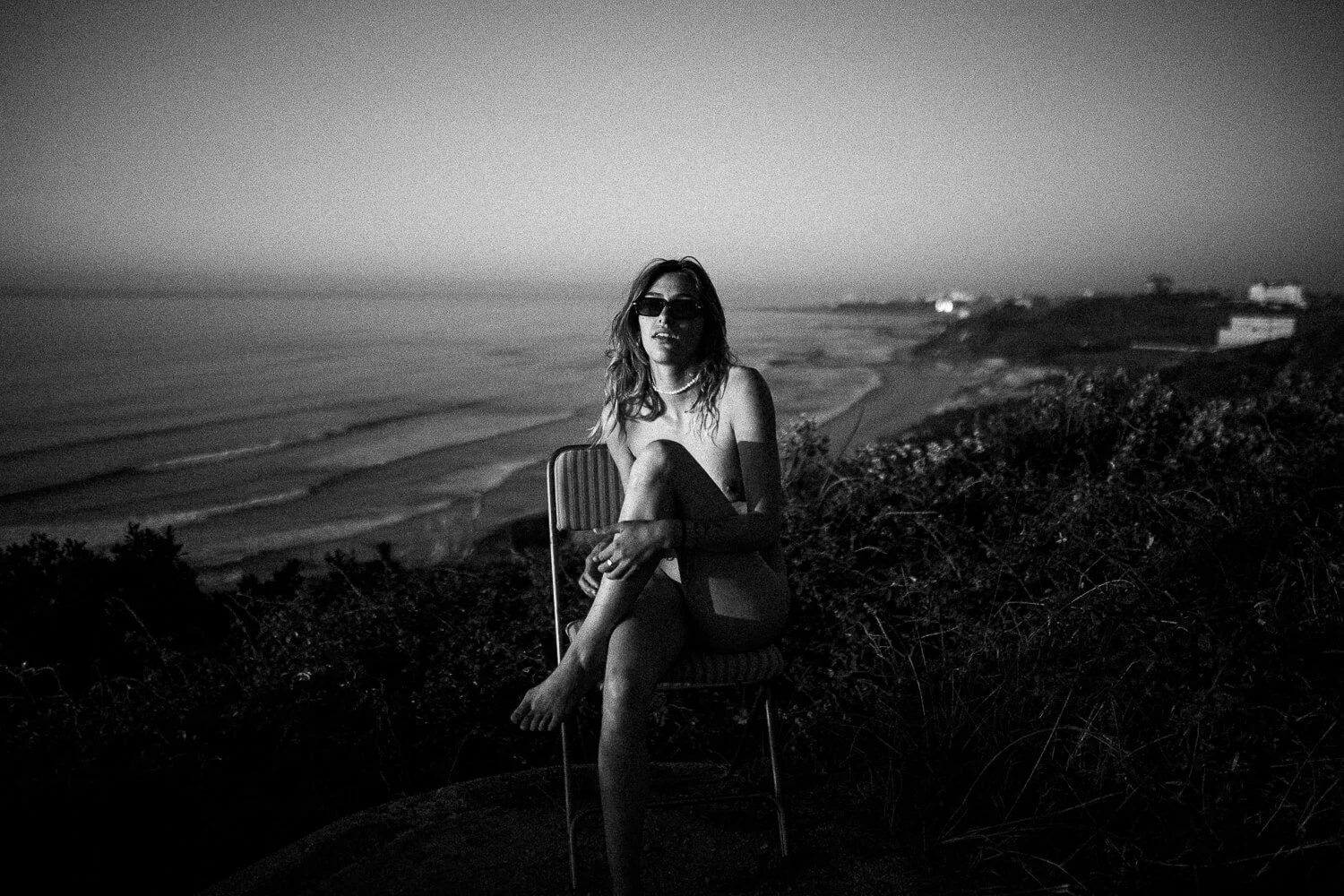 A woman sitting on a chair outdoors near the beach at dusk, wearing sunglasses and a necklace, with the ocean and shoreline in the background in black and white.