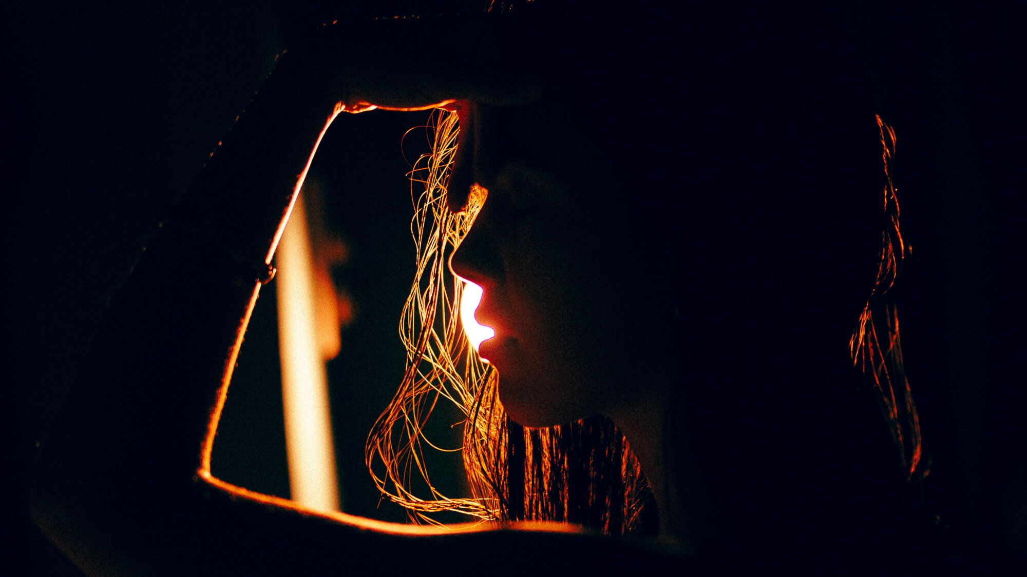 Silhouette d'une femme avec des cheveux longs, éclairée par une lumière chaude, regardant par une ouverture ou un cadre, dans un environnement sombre.
