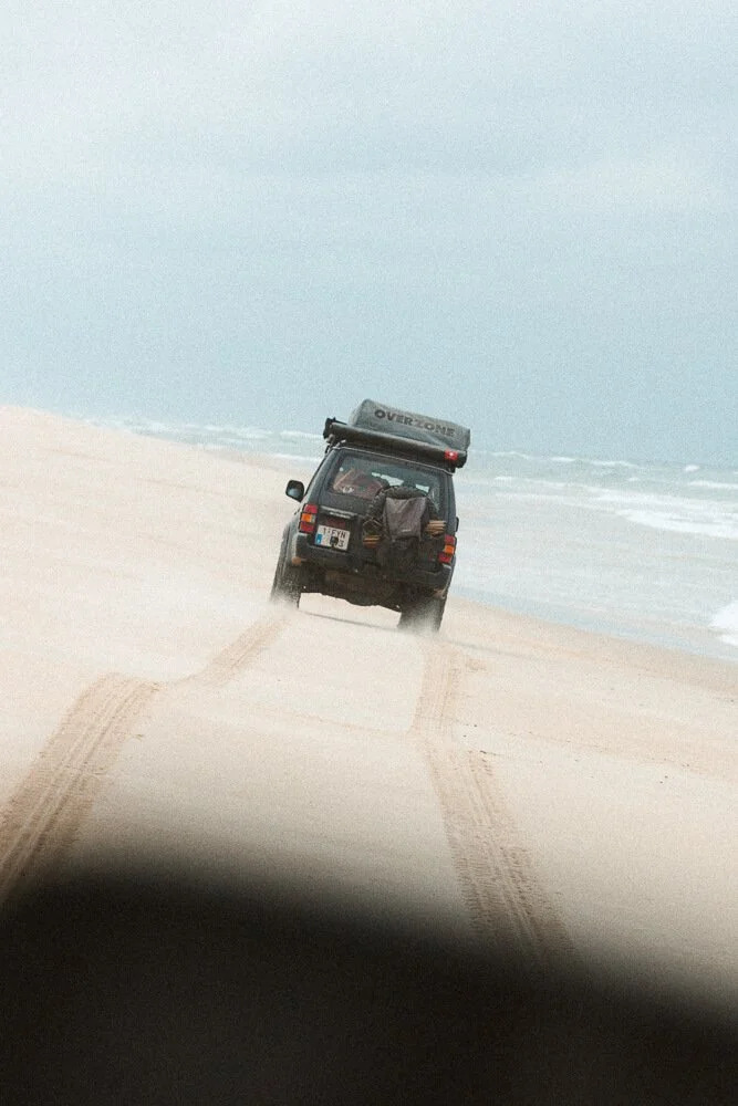 A black SUV driving on a sandy beach near the ocean with tire tracks visible and a cloudy sky.