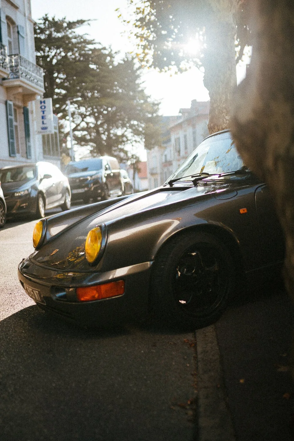 A black vintage Porsche 911 parked on the street with yellow headlights, partially obscured by a tree trunk, with other parked cars and buildings in the background on a sunny day.