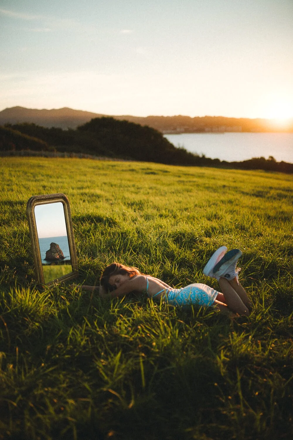 A young girl lying on green grass in a field at sunset, resting her head on her arm with her eyes closed. There is a mirror beside her reflecting a rock formation in the sea, with hills and water in the background.