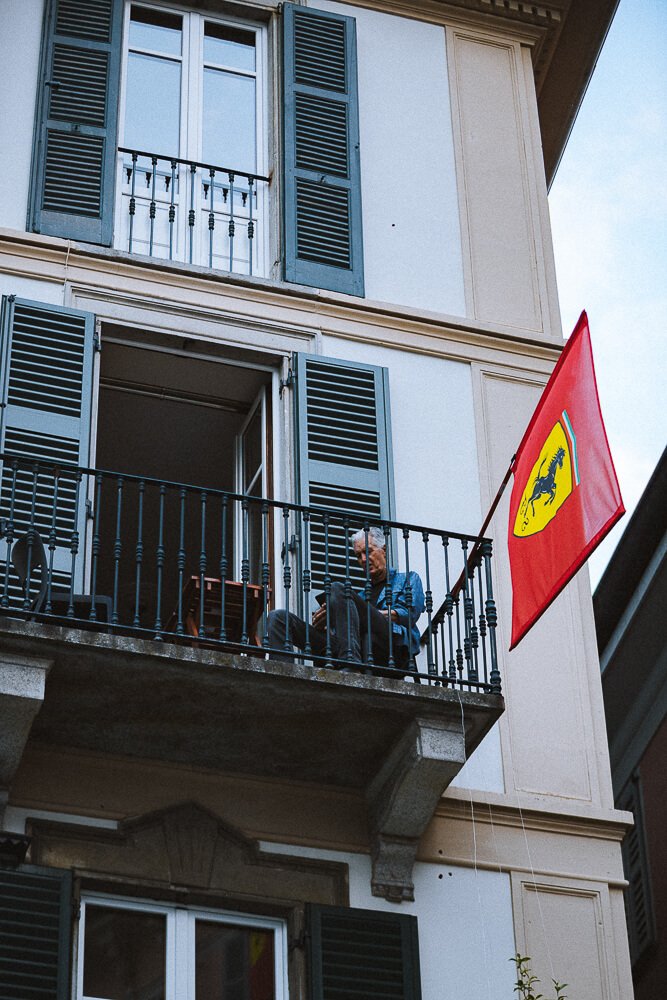A person sitting on a small balcony with green shutters, blue walls, and a red Ferrari flag.