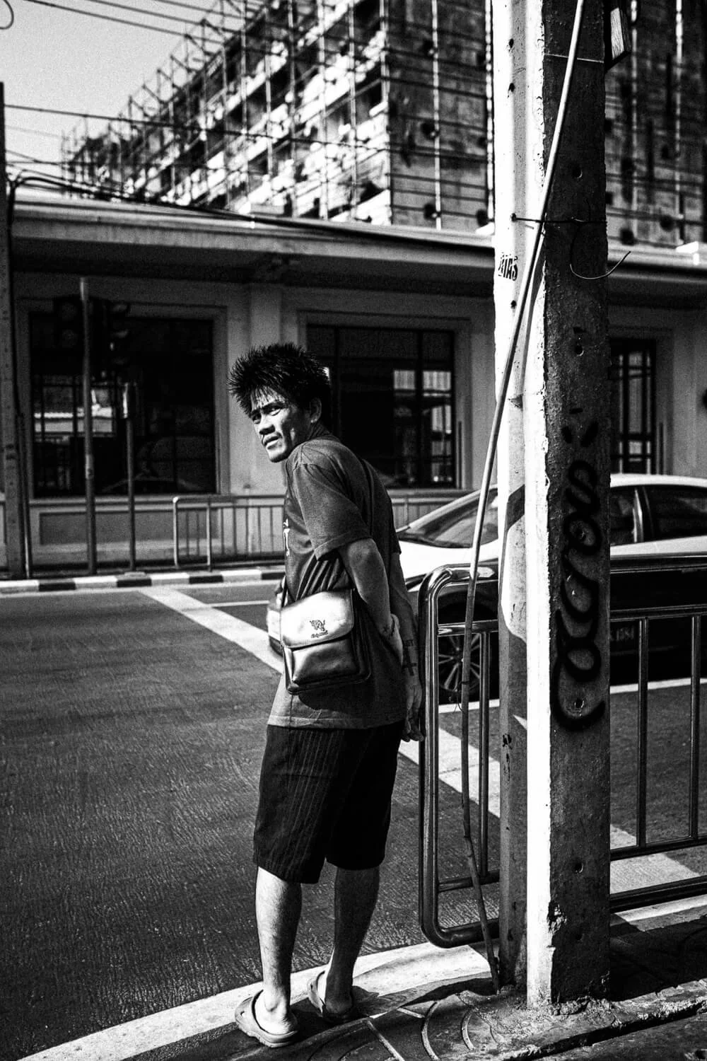 Un homme debout à un arrêt de bus dans une scène urbaine en noir et blanc, portant un sac en bandoulière.