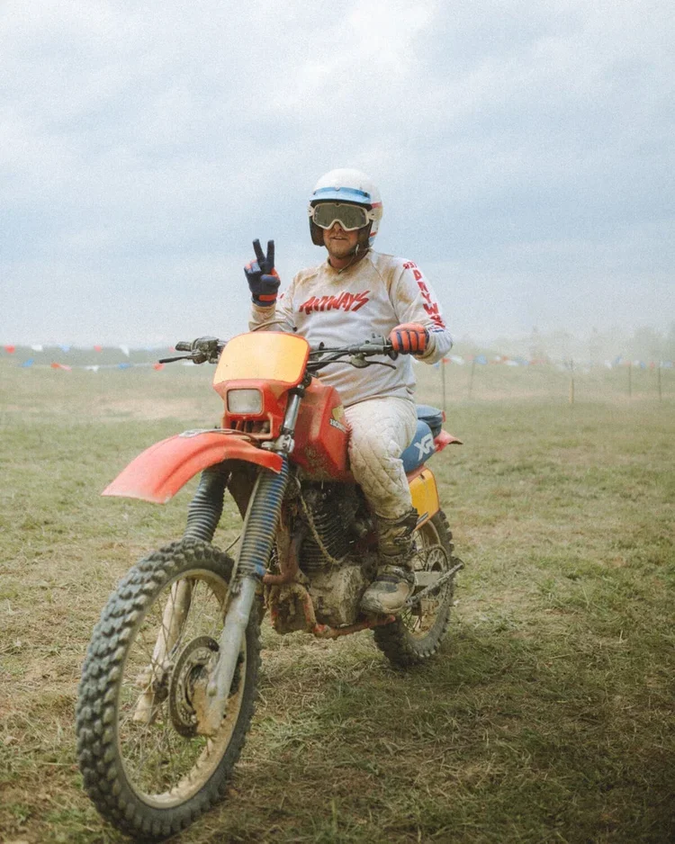 A person in motocross gear sitting on a dirt bike, making a peace sign with their right hand, on an off-road field during daytime.