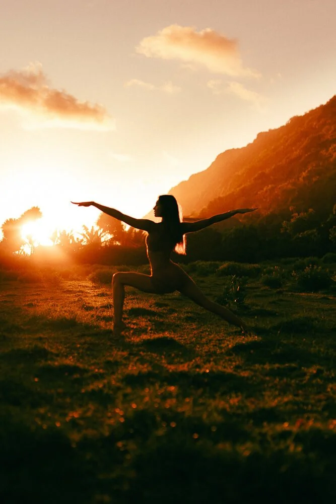 A woman practicing yoga outdoors at sunset, standing in a warrior pose with her arms extended, against a hilly landscape with trees and a colorful sky.