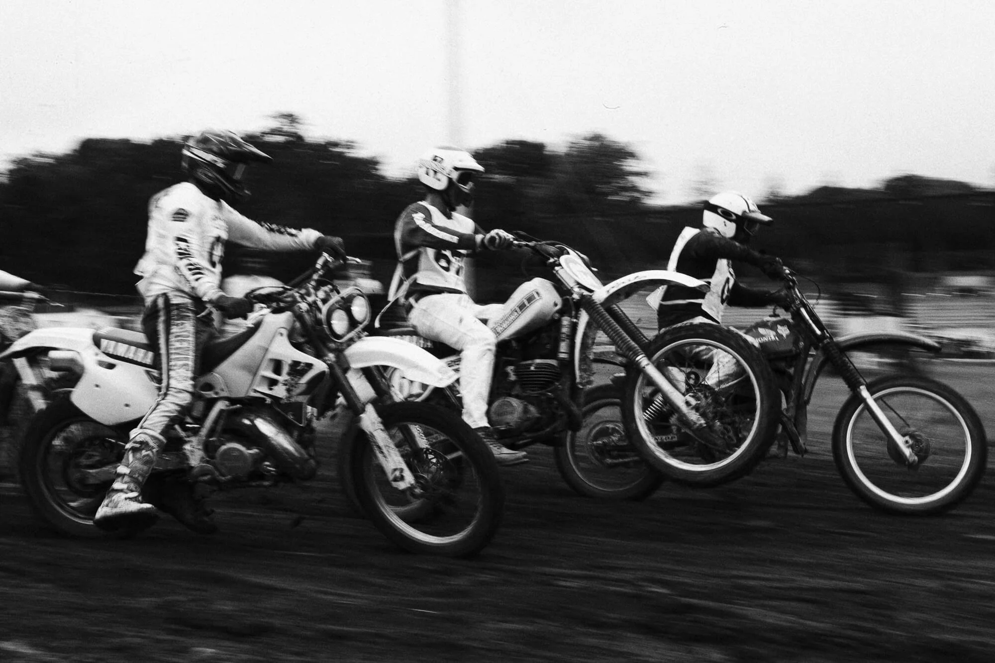 Trois motards en course, photographiés en noir et blanc, avec un mouvement flou en arrière-plan.