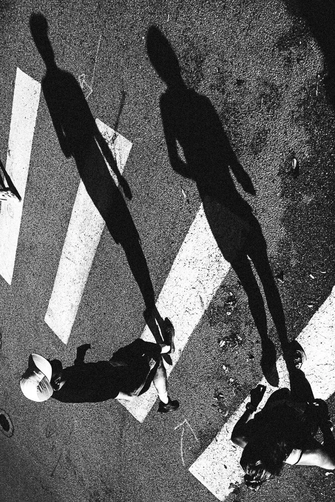 Shadow of two pedestrians crossing the street at night, cast on the pavement with white crosswalk lines visible.