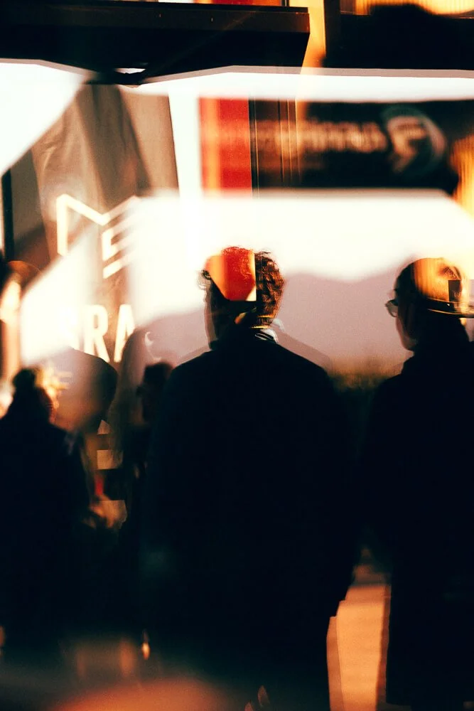 Silhouettes of people at a sunset event, with a large screen displaying a partial logo or text in the background.