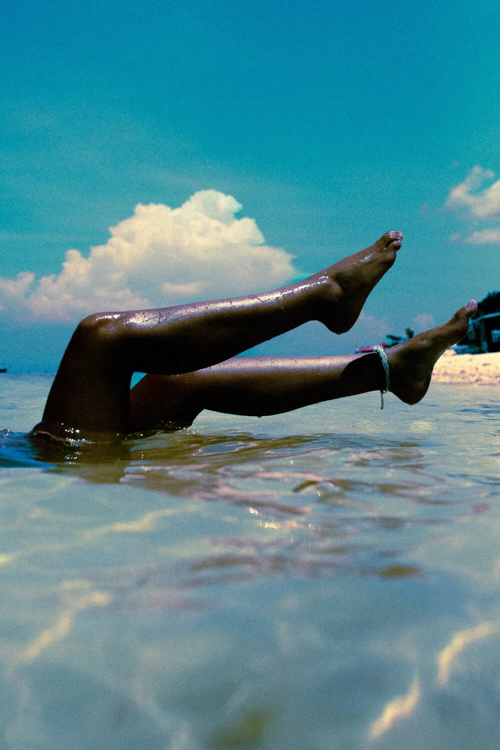 Deux jambes humaines qui sortent de l'eau dans un environnement de plage avec un ciel bleu et des nuages blancs.