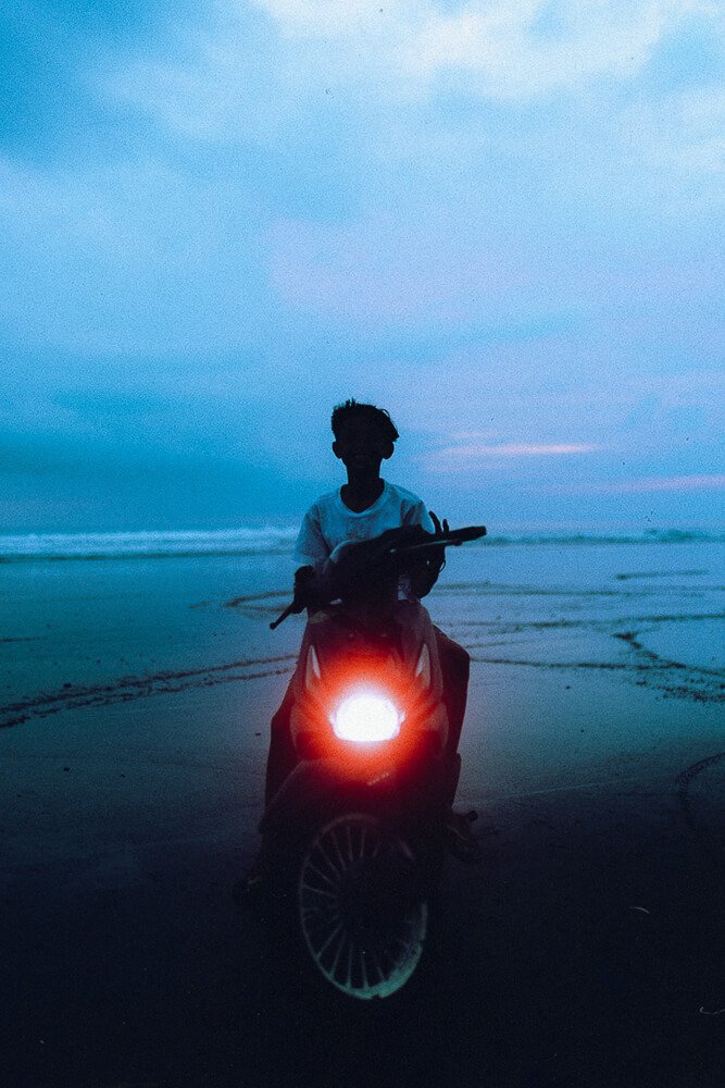 Silhouette of a person on a motorcycle with headlight on, holding a skateboard, on a beach at dusk or dawn.