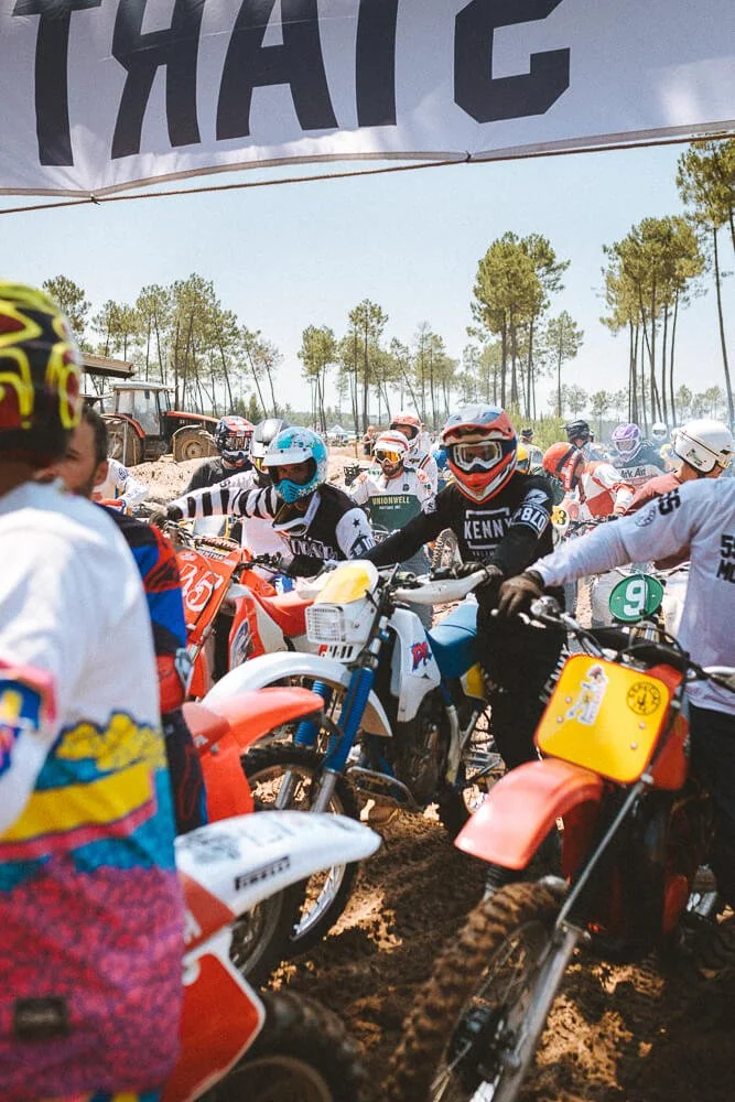Group of motocross riders wearing helmets and gear gathered on a dirt track, with a banner overhead and trees in the background.