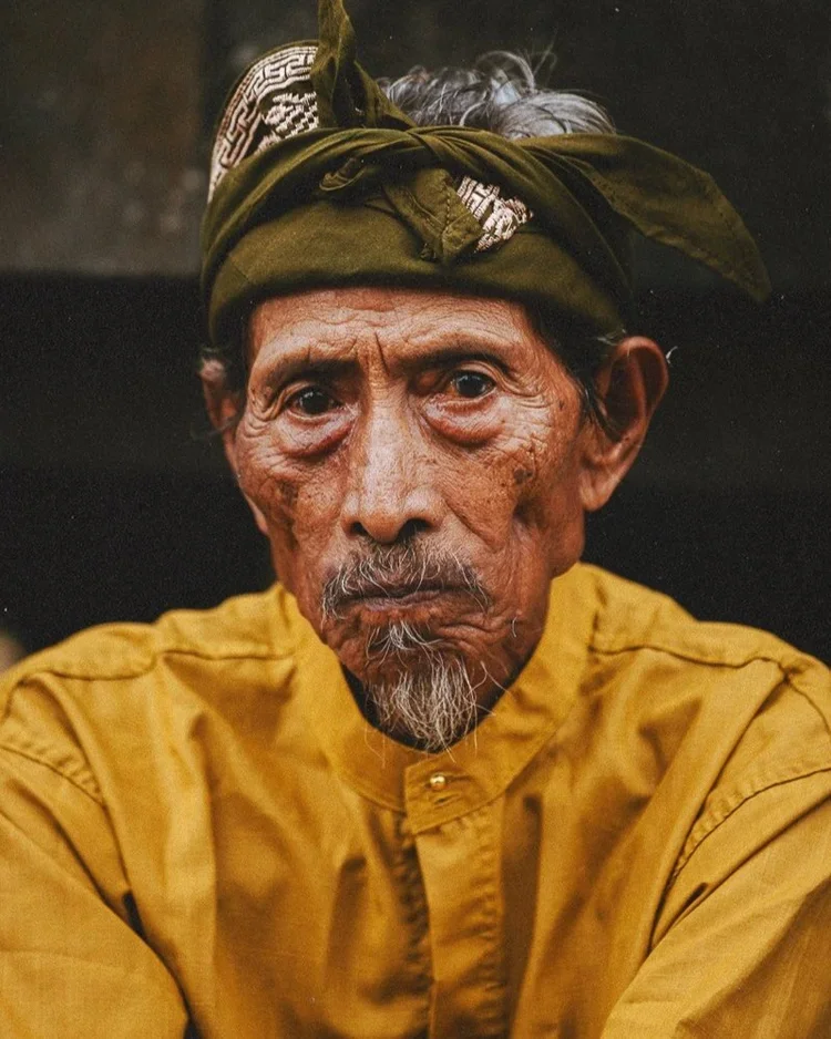 Portrait d'un homme âgé avec une peau ridée et un regard sérieux. Il porte un bandana vert sur la tête et une chemise jaune.