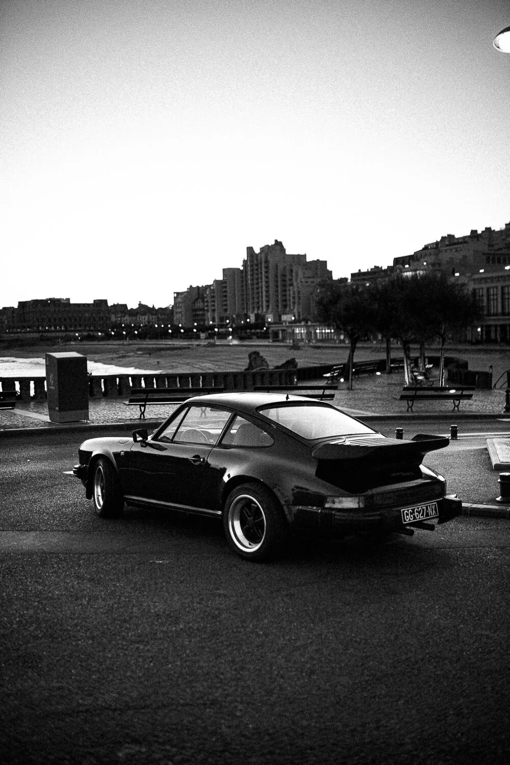 A black classic sports car parked on the street at dusk, with city buildings, trees, benches, and a beach in the background.