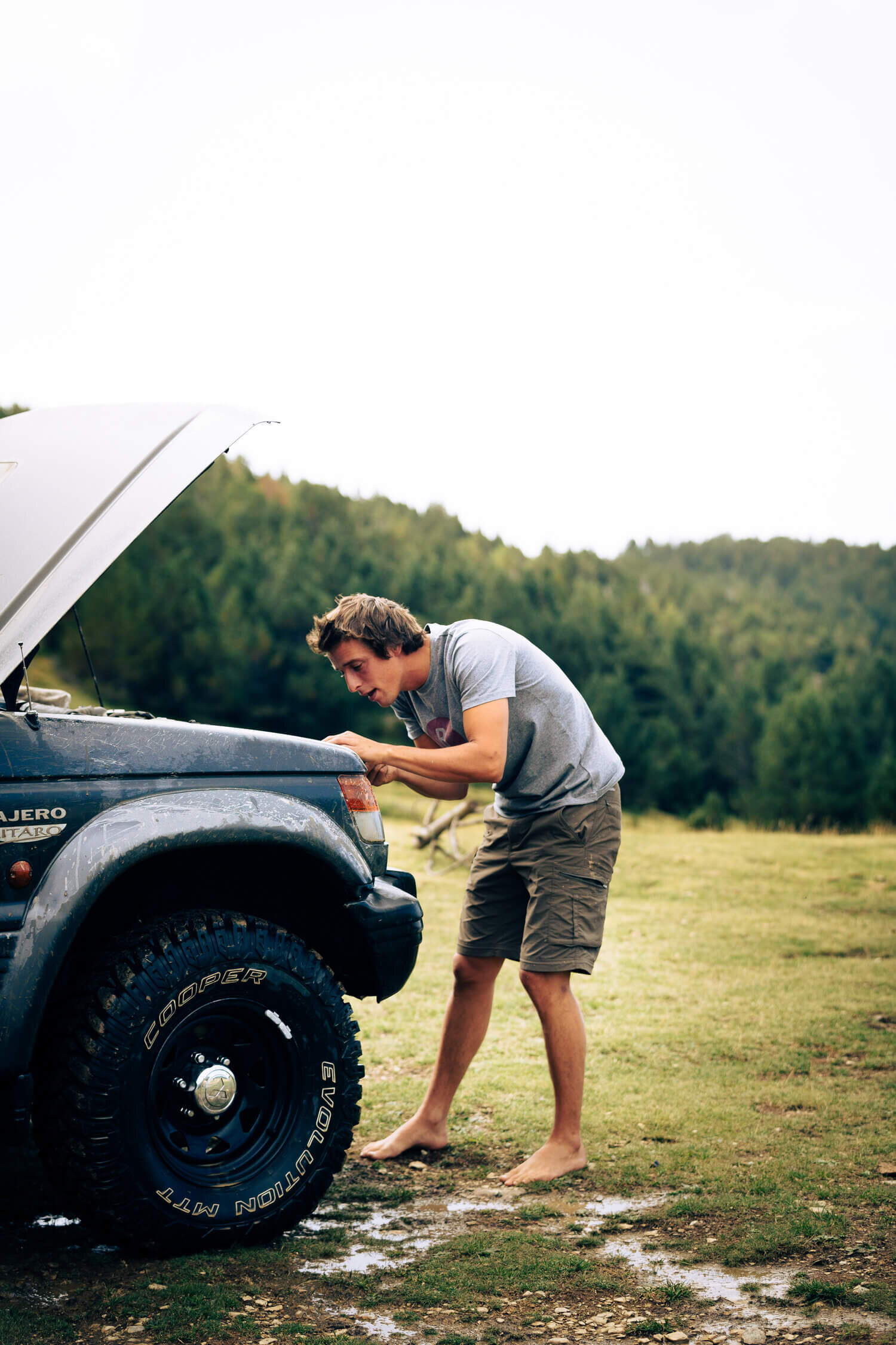 Un homme pieds nus examine le moteur d'un 4x4 avec le capot ouvert, debout sur un terrain herbeux devant une forêt.