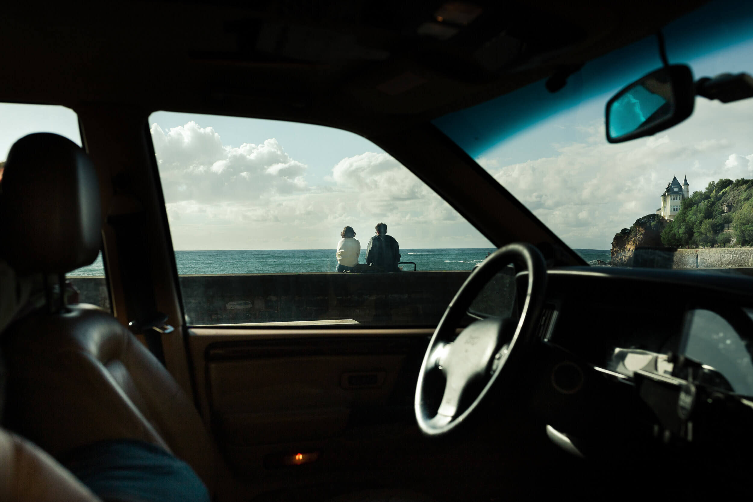 Vue depuis l'intérieur d'une voiture, deux personnes assises sur un muret regardant la mer, ciel nuageux, château distant sur une falaise.