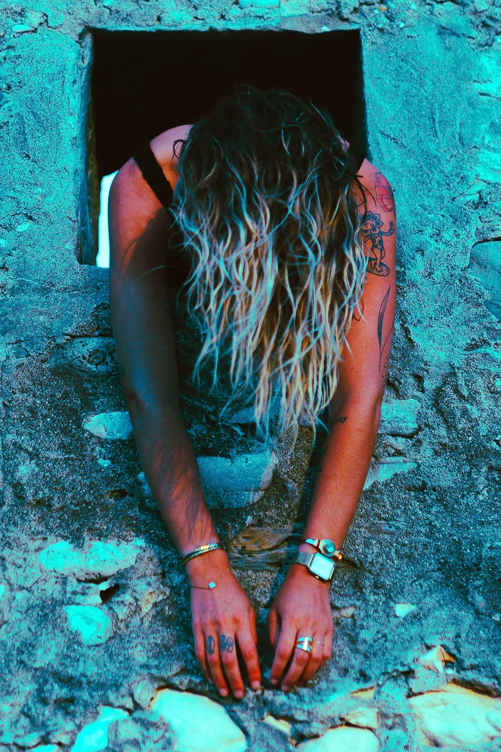A woman with long, wavy blonde hair and tattoos on her arms is emerging from a square opening in a stone wall, with her hands on the ground and head bowed down.