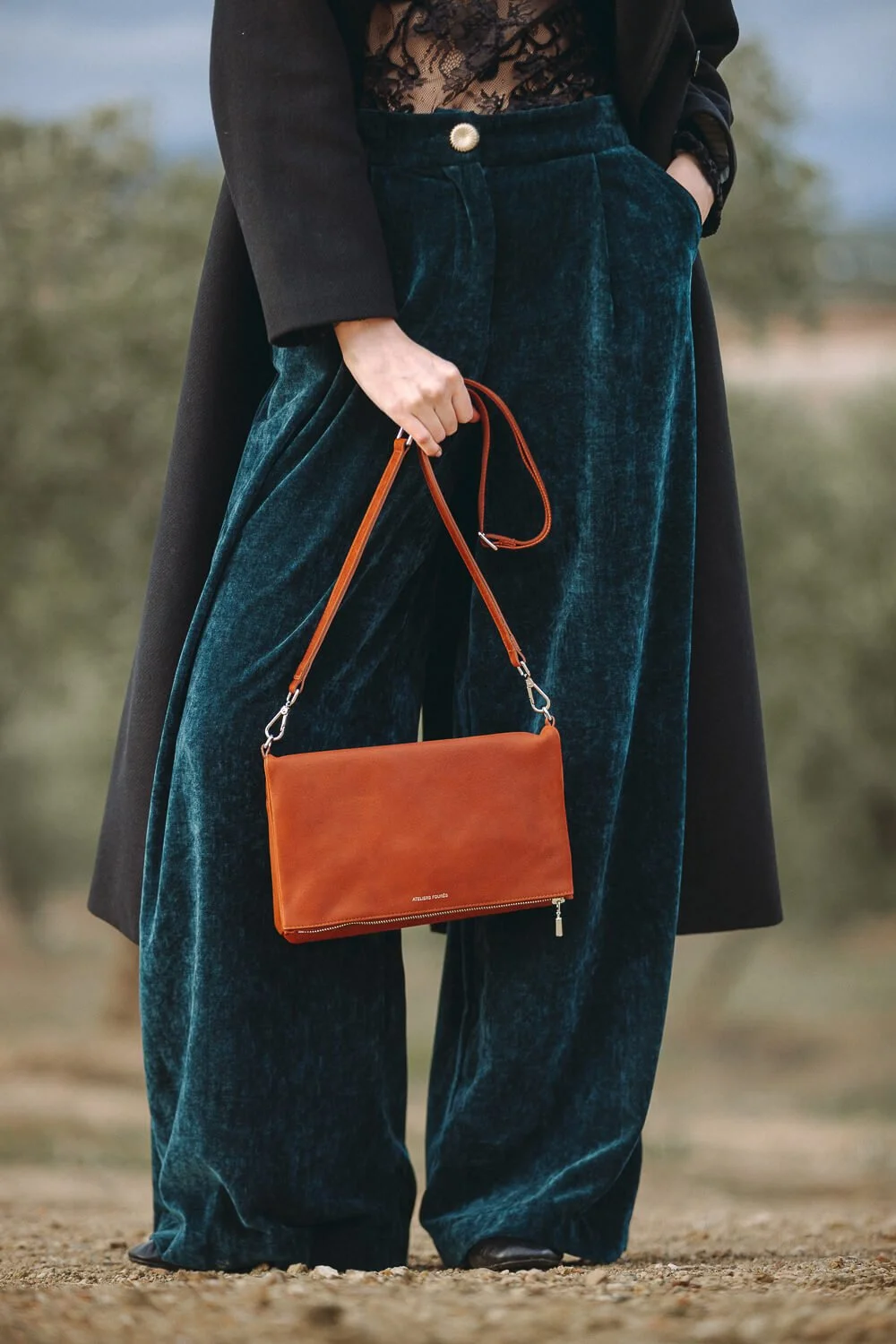 Close-up of a woman wearing wide-legged velvet pants, a lace top, and a long coat, holding an orange handbag outdoors.