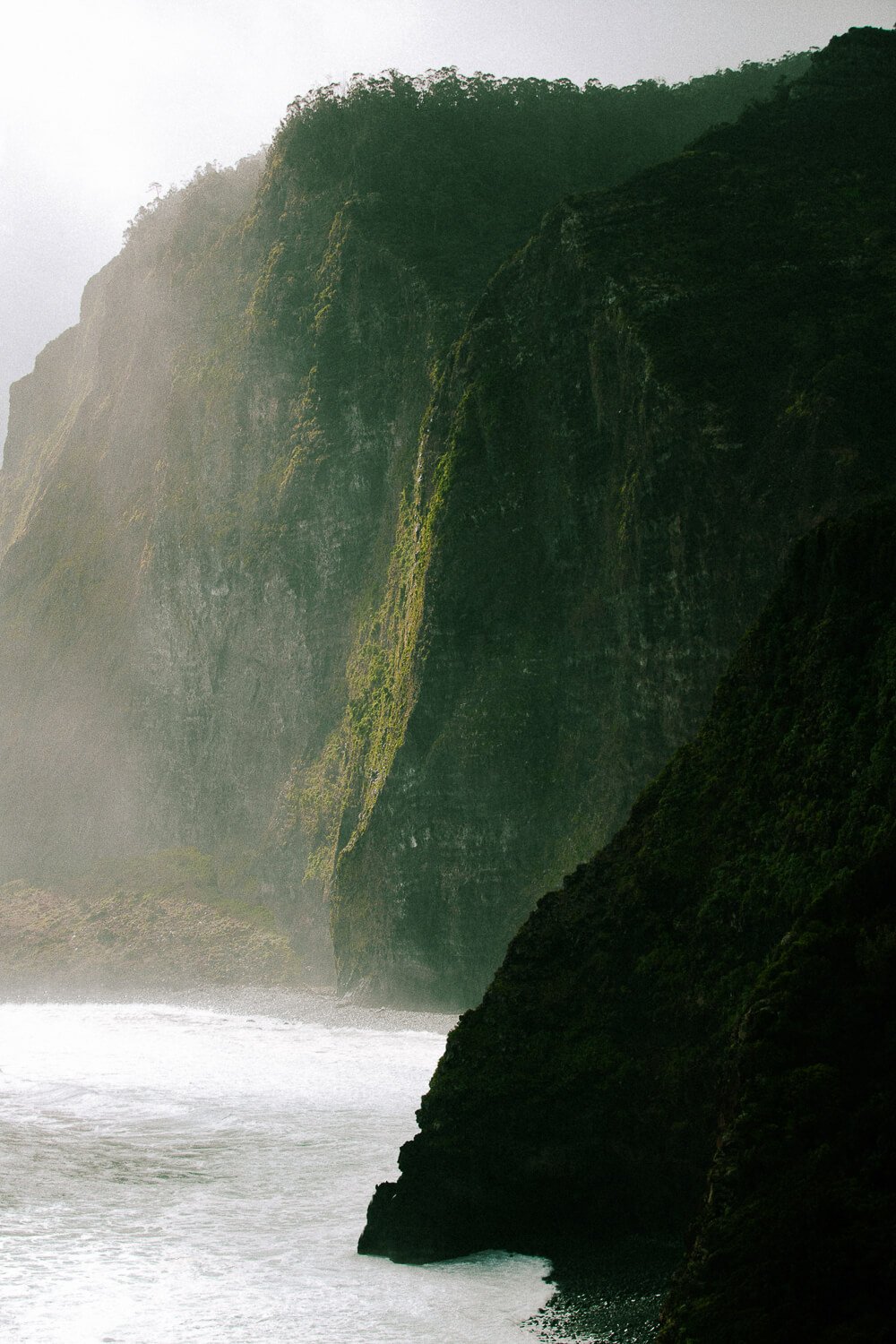 A coastal scene with towering green moss-covered cliffs, mist, and waves crashing at the base.