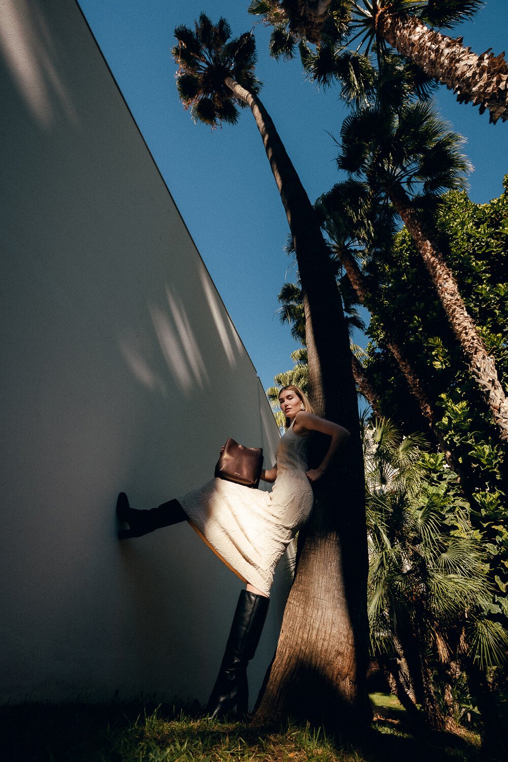 A woman in a beige dress and tall black boots leaning against a tree, holding a brown purse, with palm trees and a clear blue sky in the background.