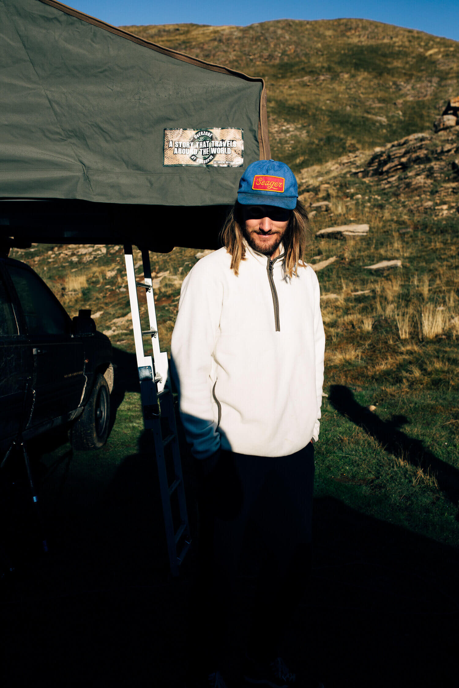 Un homme avec une casquette bleue et un pull blanc se tient à côté d'un véhicule et d'une tente de camping dans un paysage montagneux.