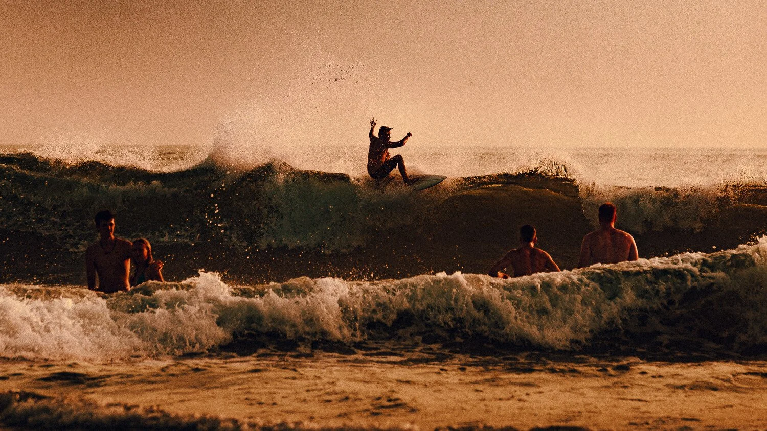 Une personne fait du surf sur une vague au coucher du soleil, avec plusieurs autres personnes dans l'eau à proximité.