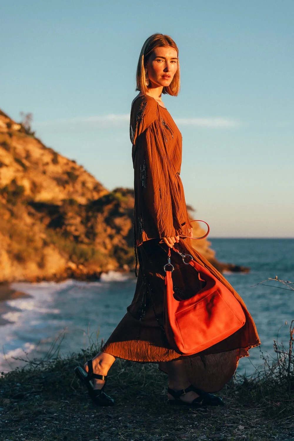 A woman standing on a hill by the ocean during sunset, wearing a long brown dress, holding a red bag, with a rocky hillside in the background.