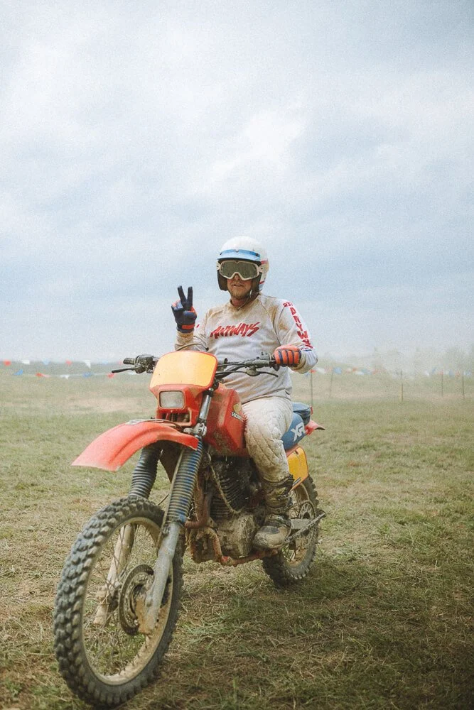 A person sitting on a dirt bike in an open field, wearing a riding helmet, goggles, and a long-sleeve shirt, making a peace sign with one hand. The sky is cloudy and there are some distant fence posts in the background.