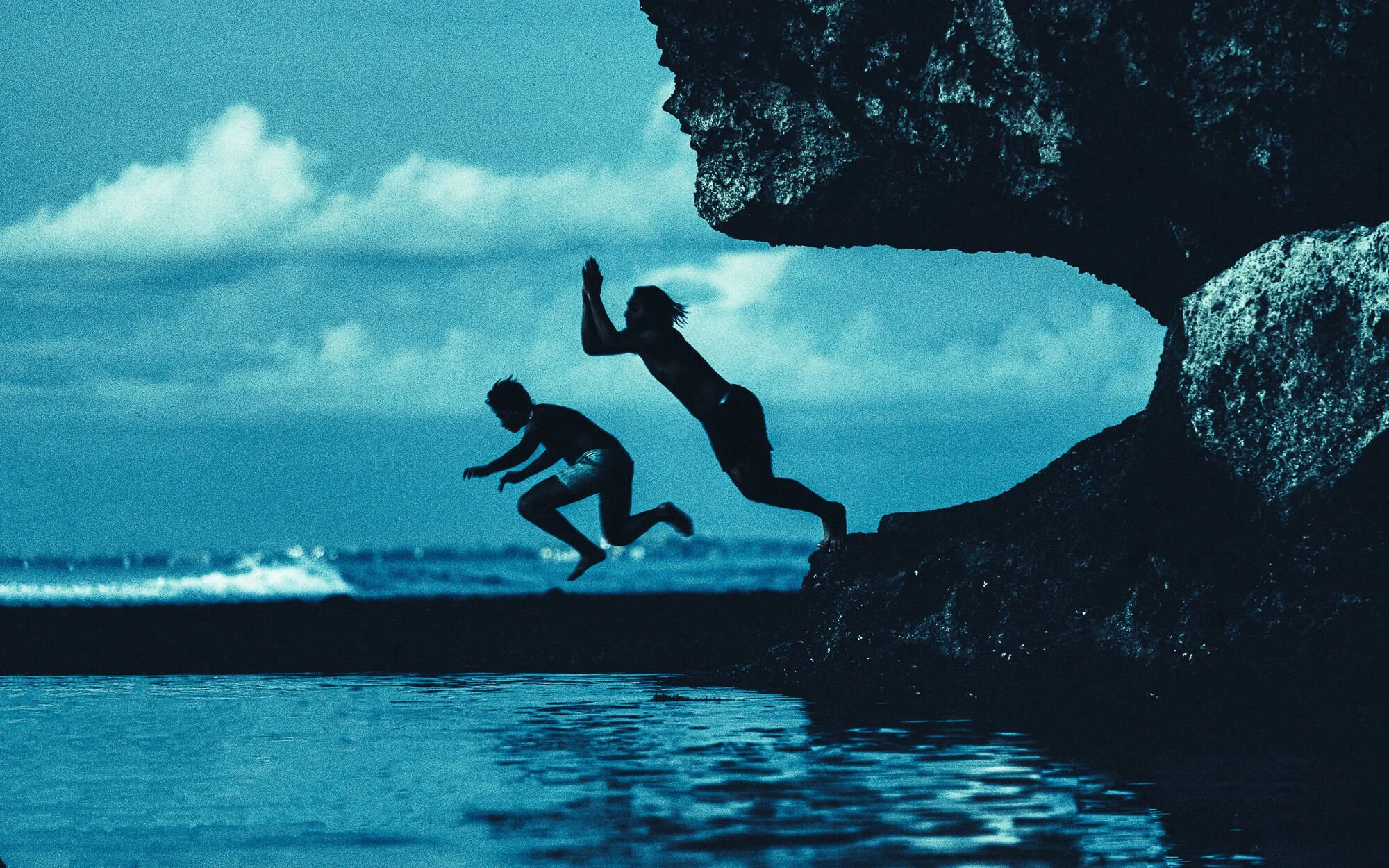 Deux personnes sautant dans l'eau depuis un rocher sur la plage, avec un ciel nuageux et la mer en arrière-plan.