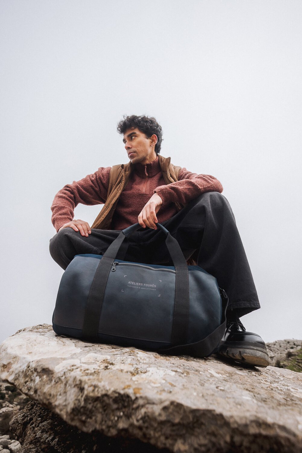 Young man sitting on a rock, with a large black duffle bag labeled 'Ateliers Fourés' in front of him, against a plain white background.
