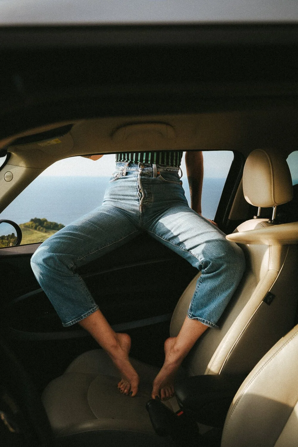 Person sitting in the backseat of a car with legs spread on the seat and head outside the car window, with a scenic landscape in the background.