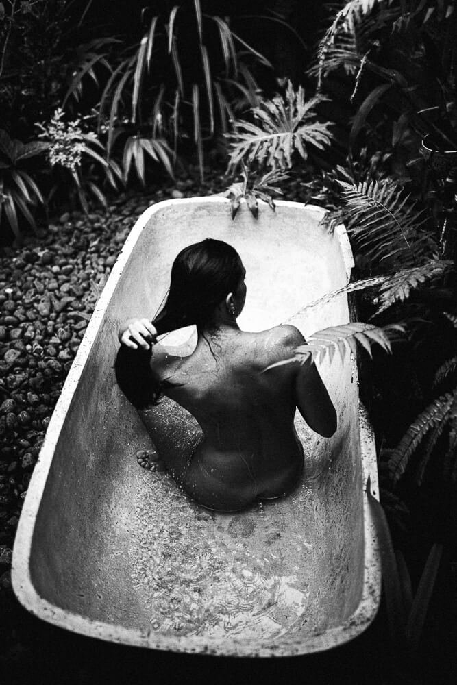 A woman sitting in an outdoor bathtub surrounded by lush plants, seen from behind, in black and white.